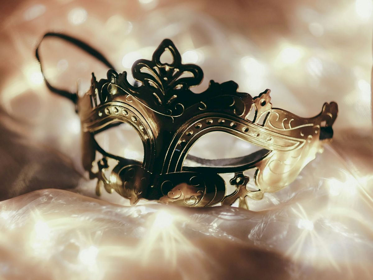 A black and gold ornate masquerade mask lies on a soft, glowing surface, surrounded by a warm, twinkling light.