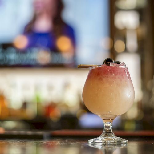 A cocktail in a frosty glass garnished with a cherry sits on a bar counter with a blurry TV screen and bar shelves in the background.