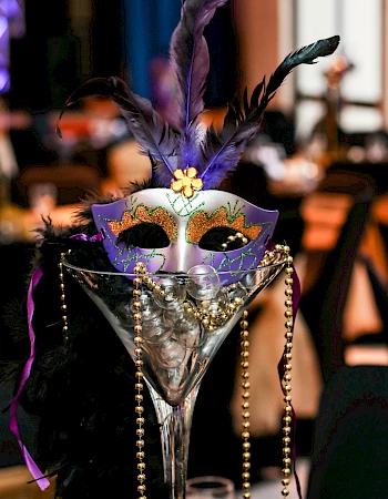 A decorative masquerade mask with feathers, gold beads, and a black feather boa are arranged in a large martini glass, likely for a celebration.