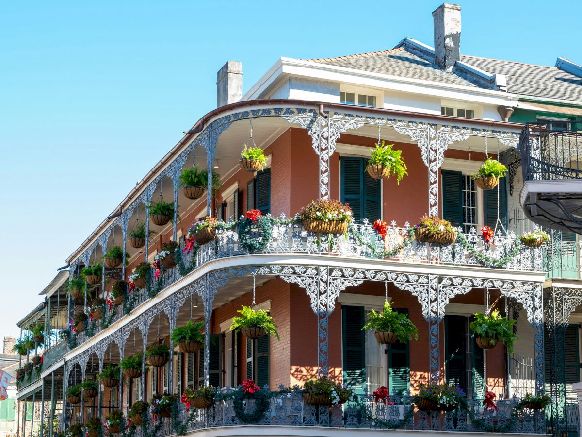 A corner building with ornate iron balconies adorned with lush greenery and flowers under a clear blue sky, possibly in a historic district.